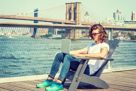 Young Hispanic American Man traveling in New York, with long hair, wearing sunglasses, white T shirt, jeans, sneakers, sitting on chair by East River, working on laptop computer. Bridges on backgroundの写真素材