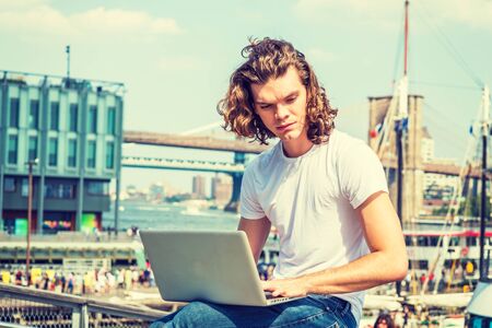 Young Hispanic American Man traveling, working in New York, with long curly hair, wearing white T shirt, sitting by East River, working on laptop computer, thinking. Bridges, boats on background.の写真素材