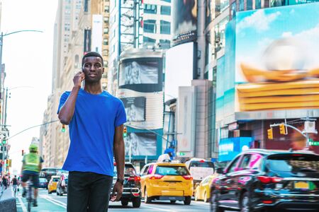 Young African American Man traveling in New York, wearing blue T shirt, walking on busy street in Times Square of Manhattan, talking on cell phone. High buildings, billboards, cars, bike on backgroundの写真素材