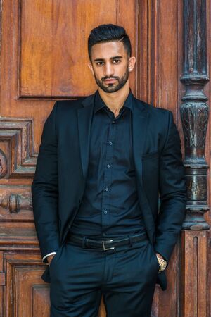 Portrait of Young Handsome East Indian American Businessman with beard in New York, wearing black suit, black shirt, hands in pockets, standing by brown old style office doorway, looking at you.の写真素材
