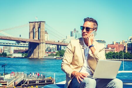 Young European Businessman traveling, working in New York, with beard, little gray hair, wearing beige blazer, sunglasses, sitting by East River, working on laptop computer, talking on cell phone.の写真素材