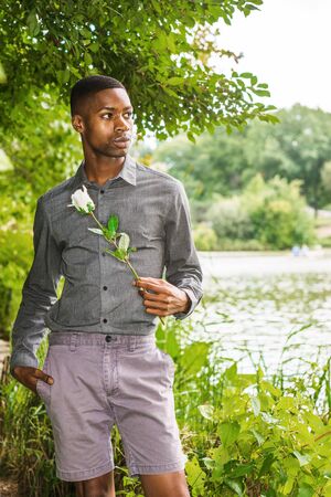 Young African American Man missing you and waiting for you at Central Park in New York. Young black man standing by lake, holding white rose, waiting, thinking, lost in thought.の写真素材