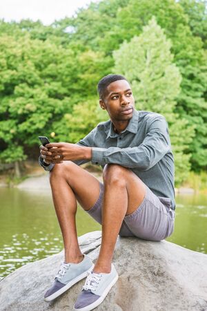 Young African American Man wearing gray long sleeves shirt, shorts, sneakers, sitting on rocks by lake, texting on cell phone, looking away, thinking.の写真素材