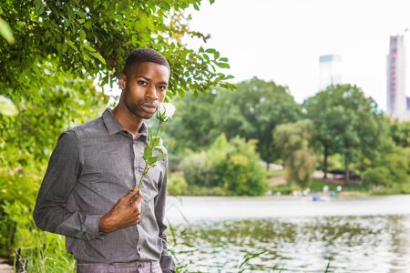 Young African American Man missing you and waiting for you at Central Park in New York. Young black man standing by lake, holding white rose, smelling, waiting, thinking.の写真素材