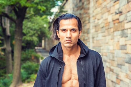 Young East Indian American Man wearing black hooded outwear, opened, showing half naked body, standing on narrow road with rock wall in New York, looking forward.の写真素材