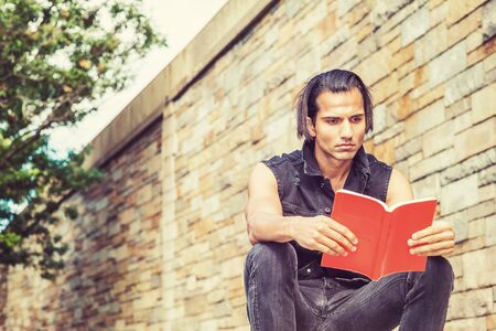 Young Modern East Indian American Man wearing black sleeveless jacket, jeans, sitting by stone wall...