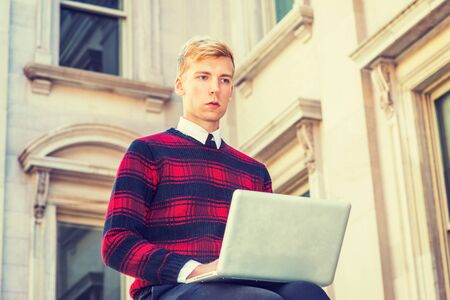 Young blonde American college student wearing patterned red, black knit sweater,  sitting outside vintage office building on campus, working on laptop computer, looking up, thinking.の写真素材