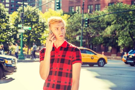 Young blonde American Man wearing red patterned Polo shirt, standing on busy street in New York, with many cars on background, under sun, looking around, calling on cell phone.の写真素材