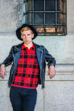 Young Man Casual Spring/Autumn Fashion in New York, wearing black leather jacket, patterned red undershirt, black hat, standing against vintage wall with window on street, looking at you.の写真素材