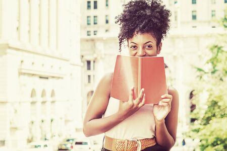 Reading outside. Young African American female college student with afro hairstyle on street by vintage style office building in New York, holding red book, covering face, smiling. Retro filtered lookの写真素材