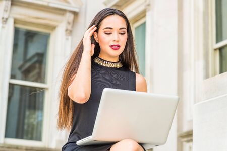 South American Female College Student studying in New York City, wearing black sleeveless dress, sitting outside office building on campus, working on laptop computer, raising arm, hand touching hair.の写真素材