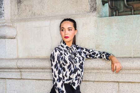 Portrait of Young South American Female College Student in New York City. Young Beautiful Hispanic Woman wearing long sleeve patterned shirt, standing against wall on campus, looking away, thinking.の写真素材