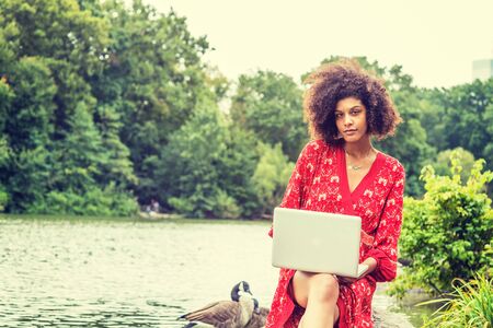 Young African American College Student with afro long curly hair, wearing red patterned dress, sitting on rocks by lake at Central Park, New York City, working on laptop computer, looking, thinking.の写真素材