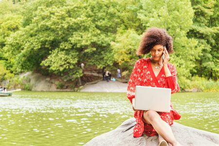 Young African American College Student with afro curly hair, wearing red patterned dress, sitting on rocks by lake at Central Park, New York City, working on laptop computer, talking on cell phone.の写真素材