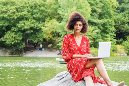 Young African American College Student with afro curly hair, wearing red patterned dress, sitting on rocks by lake at Central Park, New York City, working on laptop computer, looking up, thinking.の写真素材