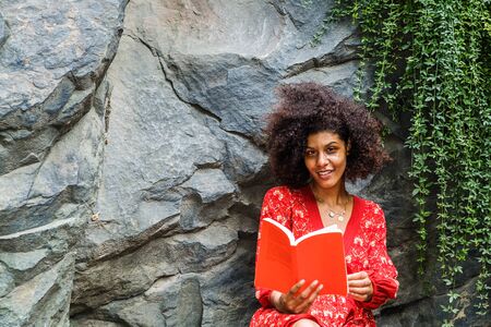 Young Mixed Race African American Woman with afro long curly hair reading red book outdoor at Central Park, New York City, wearing red flower patterned dress, sitting against rocks with green leaves.の写真素材