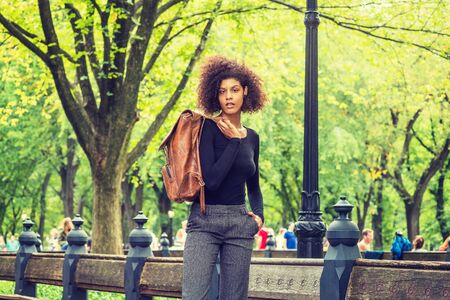 Street Fashion in New York City. Young African American Woman with afro hairstyle, dressing in black sweater, dark gray pants, carrying brown leather bag on shoulder, standing at park with green treesの写真素材