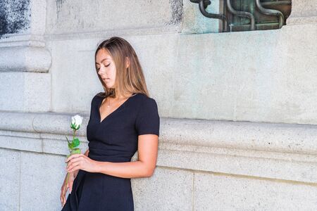I love you, waiting for you. Young East European Woman wearing black short sleeve, v neck dress, holding white rose flower, standing by wall on street in New York City, looking down, sad, thinking.の写真素材