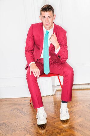 Young Businessman Fashion in New York City. 20 years old man with short hair, wearing red suit, white shirt, white sneakers, light green tie, siting on red chair by white background, looking at you.の写真素材