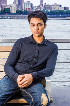 Portrait of 18 years old East Indian American teenager in New York City. Young student wearing black long sleeve shirt, broken fashionable jeans, sitting on chair by East River, relaxing, thinking.の写真素材