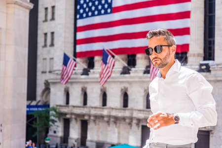 Young European Man with beard traveling in New York in summer, wearing long sleeve white shirt, sunglasses, standing outside old style office building with American flags under sun, looking forward.の写真素材
