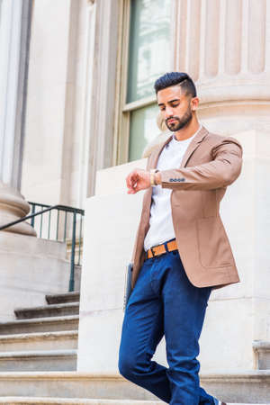 Young East Indian American Businessman with beard working in New York City, wearing brown blazer, blue pants, raising arm, looking down at wristwatch, walking down stairs from office building.の写真素材
