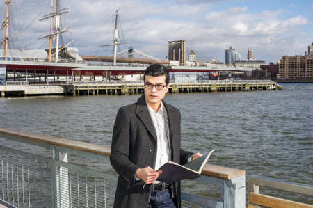 A young handsome scholar  is holding a book and standing on a river bank, looking around surrounding environment and thinkingの写真素材