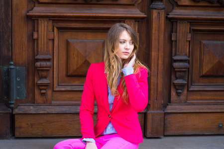 Dressing in a pink blazer, light blue under shirt and pink pants, a blonde teenage girl is casually sitting on door steps,  looking away.の写真素材