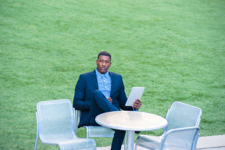 A young handsome black man is sitting on a chair by a green lawn and reading at a small computer.の写真素材