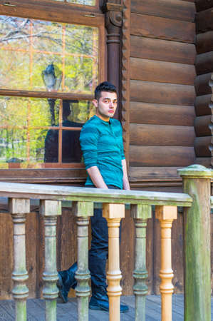 A young handsome guy is standing by a window outside a wooden house, thoughtfully looking at you. Spring greens are reflected on the window glass.の写真素材