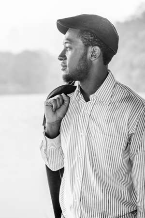Rainy Day, a little foggy, wet feel. Wearing a long sleeves shirt, a ivy cap, taking off a jacket on his shoulder,  a young handsome black man is standing by a foggy lake, confidently looking forward.の写真素材