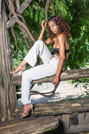 Dressing in black bra, white pants and sandals, a young, pretty girl with curly long brown hair is sitting under the shade of a wood area in summer, relaxing and thinking.の写真素材