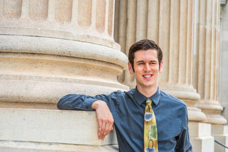 Dressing in a blue shirt, a colorful tie, a young college student is standing outside an office building, thinking.の写真素材