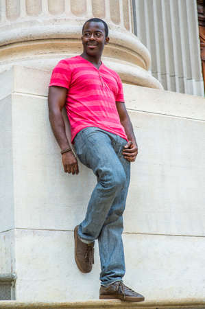 Wearing a red, pink stripe Henley V Neck T shirt,  gray pants, leather sneakers, a young black guy is standing against the base of a column, smiling and waiting for you.の写真素材