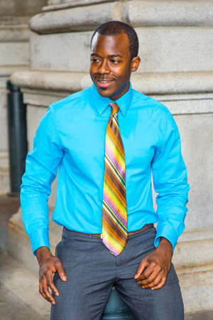 Dressing in a light blue shirt, a colorful pattern tie, a young black businessman is smiling, sitting outside to take a break.の写真素材