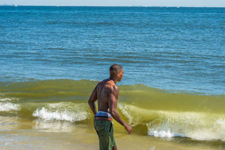 A well built young sexy black guy, half naked, whole body wet,  is walking into water of ocean, huge rolling splash waves.の写真素材