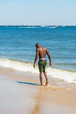 Looking down, half naked, whole body wet, a young black guy is walking on the beach. The horizontal is a big city outline, small boats on the background.の写真素材