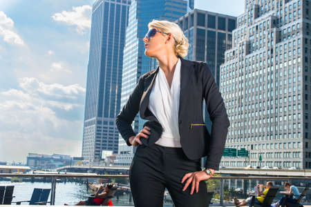 Dressing in white underclothes and a black suit unbuttoned, wearing sunglasses, a young pretty blonde businesswoman is standing in the front of a business district and looking forward.の写真素材