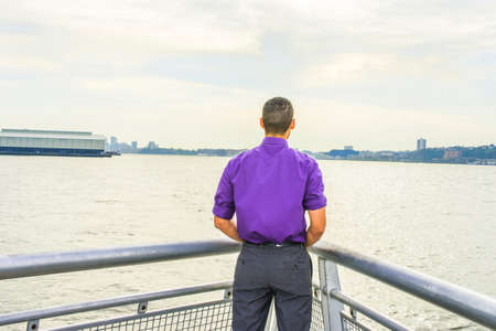 Young Man facing water in back view. Dressing in a purple shirt, gray pants, a young guy with shot curly hair is standing by a river, looking faraway, waiting for you.の写真素材