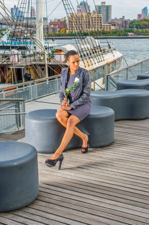 Dressing in faux fur jacket, a woolen fitted dress, open toes shoes, holding a white flower,  a young black woman is sitting on a deck, waiting for you. The background is a harbor.の写真素材