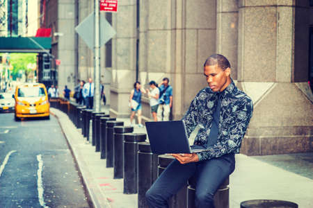 African American businessman in New York. Wearing black flower patterned shirt, tie, young guy siting on street, reading, working on laptop computer. Yellow cab, people on background.の写真素材