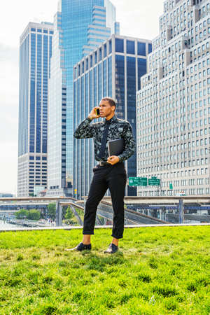African American businessman working in New York. Wearing black flower patterned shirt, tie, holding laptop computer, young  guy standing in front of business district, talking on his mobile phone.の写真素材