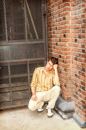 Concept of lonely man thinking about love. Wearing yellow patterned shirt, pants, sneakers, a young guy squatting at corner on street in New York, looking down, sad, thinking after busy working day.の写真素材