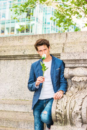 European college student seeking love in New York. Wearing blue blazer, jeans, a young guy standing at corner against wall on campus, holding white rose, smelling, thinking about you.の写真素材