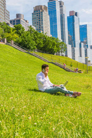 Way to Success. Wearing white shirt, jeans, sneakers,  a young college student sitting on green lawn with stairs to business district in New York, working on laptop computer, talking on phone.の写真素材