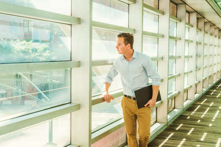 Businessman traveling, working in New York. Wearing white shirt, yellow pants, holding laptop computer, a young teacher standing by glass wall on campus, looking outside, full of sunshine, thinking.の写真素材