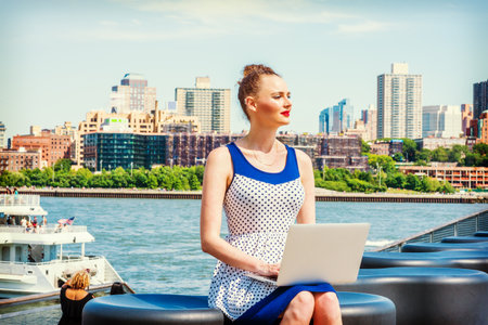 American businesswoman traveling, working in New York. Wearing blue, white polkadot dress, a young sexy lady sitting on bench at harbor, working on laptop computer, reading, thinking. Filtered effectの写真素材