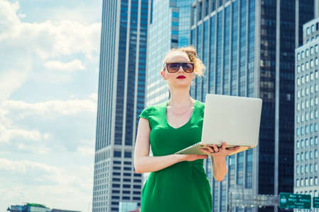 American businesswoman traveling, working in New York. Wearing green dress, sunglasses, a young sexy lady standing in business district, reading, working on laptop computer. filtered effect.の写真素材
