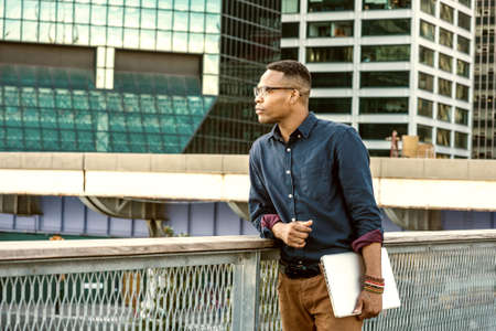 African American college student studying in New York. Wearing blue shirt, glasses, bracelets, holding laptop computer, a young black man standing in business district with high buildings, thinking.の写真素材