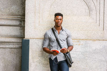 Modern College Student in New York. Wearing gray shirt, wristwatch, carrying shoulder leather bag, an African American guy standing by wall on street, listening music with earphone, reading red book.の写真素材